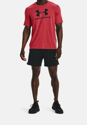 Mies, jolla on päällään punainen Under Armour -t-paita, mustat shortsit ja mustat urheilukengät, seisoo vaaleaa tasaista taustaa vasten.