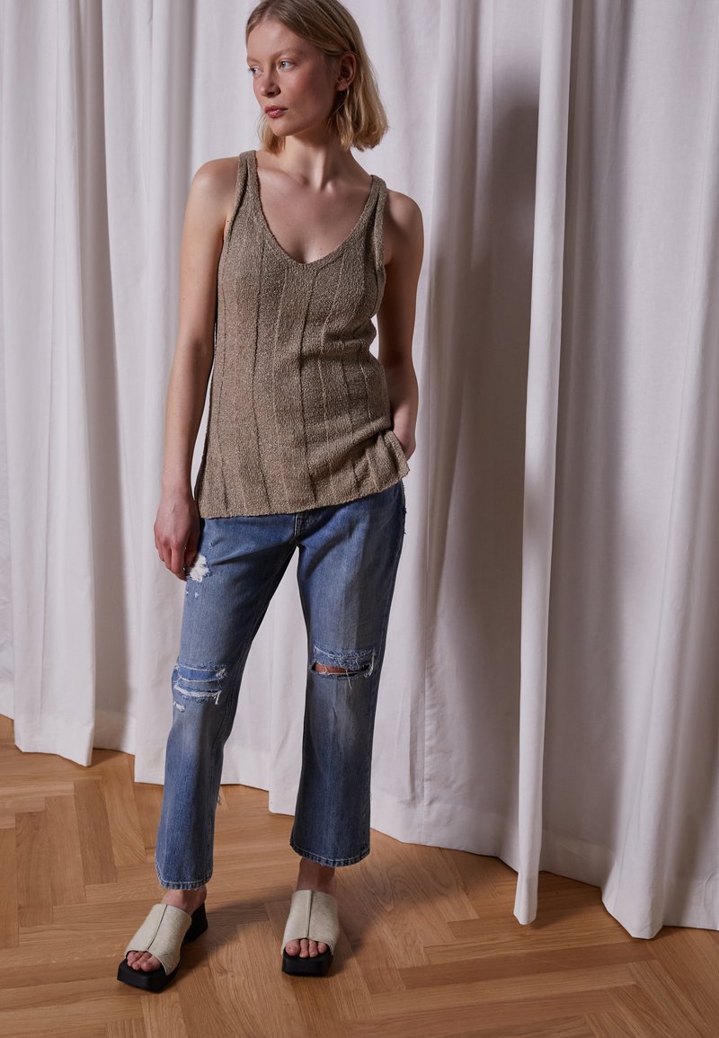 Top de malha bege com decote em V, combinado com jeans claros rasgados. Modelo usa sandálias de dedo em creme com solas pretas sobre um chão de madeira.