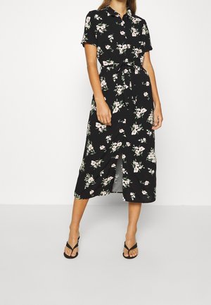 Kobieta ubrana w czarną sukienkę midi w białe kwiaty, z krótkimi rękawami, kołnierzykiem, guzikami z przodu, przewiązaną w talii i czarnymi sandałami.
