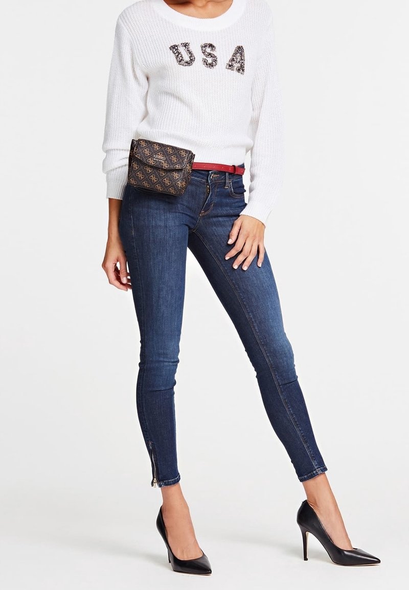 Pull en tricot blanc avec "USA" en sequins, jean skinny bleu foncé avec des zips aux poignets, ceinture rouge et escarpins noirs. Sac bandoulière marron à motifs.