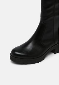 Bottines noires en cuir, mi-mollet, avec bout rond, panneaux latéraux élastiques et semelle épaisse sur fond blanc.