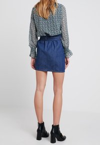 Mini-jupe en jean avec une taille ceinturée, associée à un chemisier à manches longues à motif. Des bottines noires complètent la tenue.