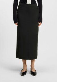Jupe crayon noire taille haute en tissu lisse ; dotée de coutures minimales et d'une poche fente, associée à des chaussures noires à bout pointu.
