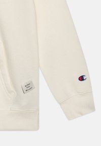 Kremfarget hettegenser med teksturert stoff, en liten etikett med teksten "Be Your Own" og et logo-lapp på ermen, med broderte detaljer.