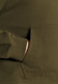 Sudadera verde oliva con puños acanalados y bolsillo tipo canguro, fabricada con una tela suave y texturizada y con detalles de costura en contraste.