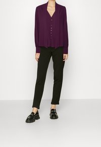 Kobieta w fioletowej bluzce na guziki, czarnych prostych spodniach z mankietami i czarnych masywnych loafersach na jednolitym białym tle.