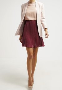 Jasnoróżowa marynarka na jasnej bluzce, połączona z bordową plisowaną spódnicą. Gładkie tekstury i neutralne szpilki dopełniają stylizację.