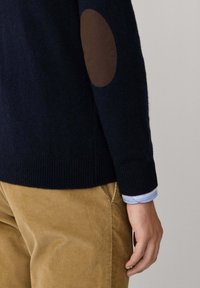 Marineblauer Pullover mit strukturiertem, geripptem Saum und Bündchen, ausgestattet mit einem braunen Wildleder-Patch an den Ellbogen. Kombiniert mit beigen Cordhosen.