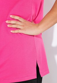 Růžový top s krátkým rukávem, hladkou texturou, bočními rozparky a volným střihem, kombinovaný s černými kalhotami se svislým pruhem.