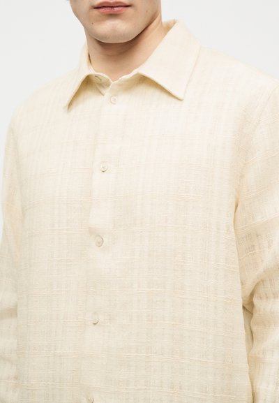 Άνδρας που φοράει μια εκρού υφασμάτινη πουκαμίσα με κουμπιά και κλασικό γιακά, εμφανιζόμενος από το στήθος και πάνω, με απλό φόντο.