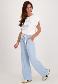 Vrouw in een witte top met opgerolde mouwen en lichtblauwe broek met koord, staand met één hand in de zak en met witte sneakers.