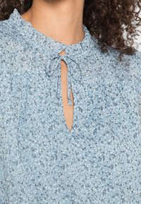 Blusa azzurro chiaro con motivo floreale, scollatura arricciata con rifiniture a volant, chiusura a cravatta e apertura a chiave parziale.