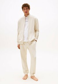 Veste zippée de couleur crème et joggers assortis en tissu doux. La veste est dotée de poches latérales et d'un détail textuel sur la manche.