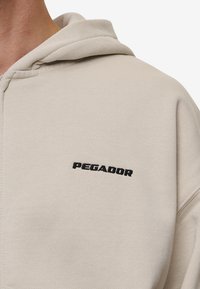 Lys beige hoodie lavet af blødt stof, med et sort "PEGADOR" logo broderet på den øverste brystdel. Indeholder en hætte med snore.