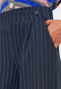 Ναυτικά παντελόνια με ρίγες και πλισέ μπροστά, με πλαϊνές τσέπες. Διαθέτει σκούρο κουμπί και διακριτική υφή στο ύφασμα.