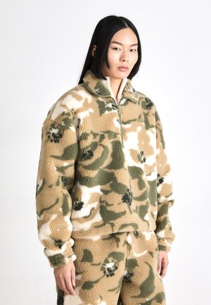 Pull en polaire avec un col zippé, arborant un motif de camouflage floral tan, vert et blanc. Texture douce et coupe décontractée.