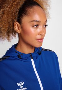 Felpa con zip blu e collo alto, accessori neri e dettagli bianchi. Presenta un tessuto testurizzato con un logo sul lato sinistro del petto.