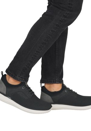 Baskets athlétiques noires et grises avec des semelles blanches portées avec un jean noir ajusté, pieds légèrement croisés en milieu de pas.