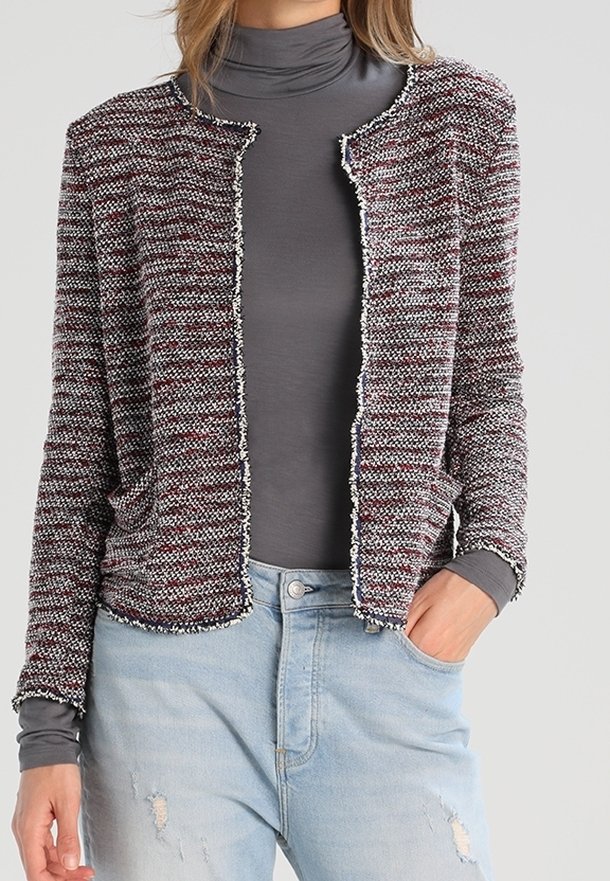 Veste en maille bordeaux et grise avec un motif texturé, devant ouvert, sans fermetures, et bordure blanche. Portée sur un col roulé gris et un jean bleu.