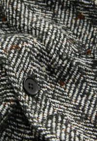 Textil s šedobielym houndstooth vzorom s textúrovanou väzbou, ktorý má ako výrazný detail čierny gombík.