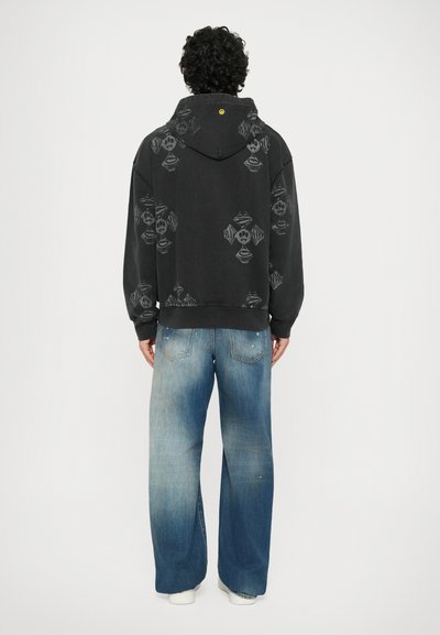 Felpa nera con cappuccio, caratterizzata da un motivo ripetuto grigio. Abbinata a jeans a gamba larga in denim chiaro, con una sfumatura che passa dal scuro al chiaro.