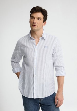 Uomo che indossa una camicia bianca con sottili righe verticali blu e maniche rimboccate, abbinata a jeans blu, guarda leggermente a sinistra.
