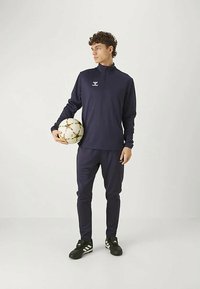 Jūras spēku apmācības treniņtērps ar rāvējslēdzēja apkakli, slim fit biksēm un melnajām futbola kurpēm. Spēlētājs tur daudzkrāsainu futbola bumbu.