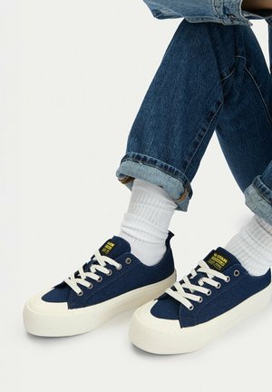 Person trägt hochgekrempelte blaue Jeans, weiße Socken und marineblaue G-Star Canvas-Sneakers mit weißen Schnürsenkeln und dicken Sohlen.