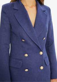 Blazer croisé bleu marine en tissu texturé, avec boutons dorés, revers en pointe et poches latérales.
