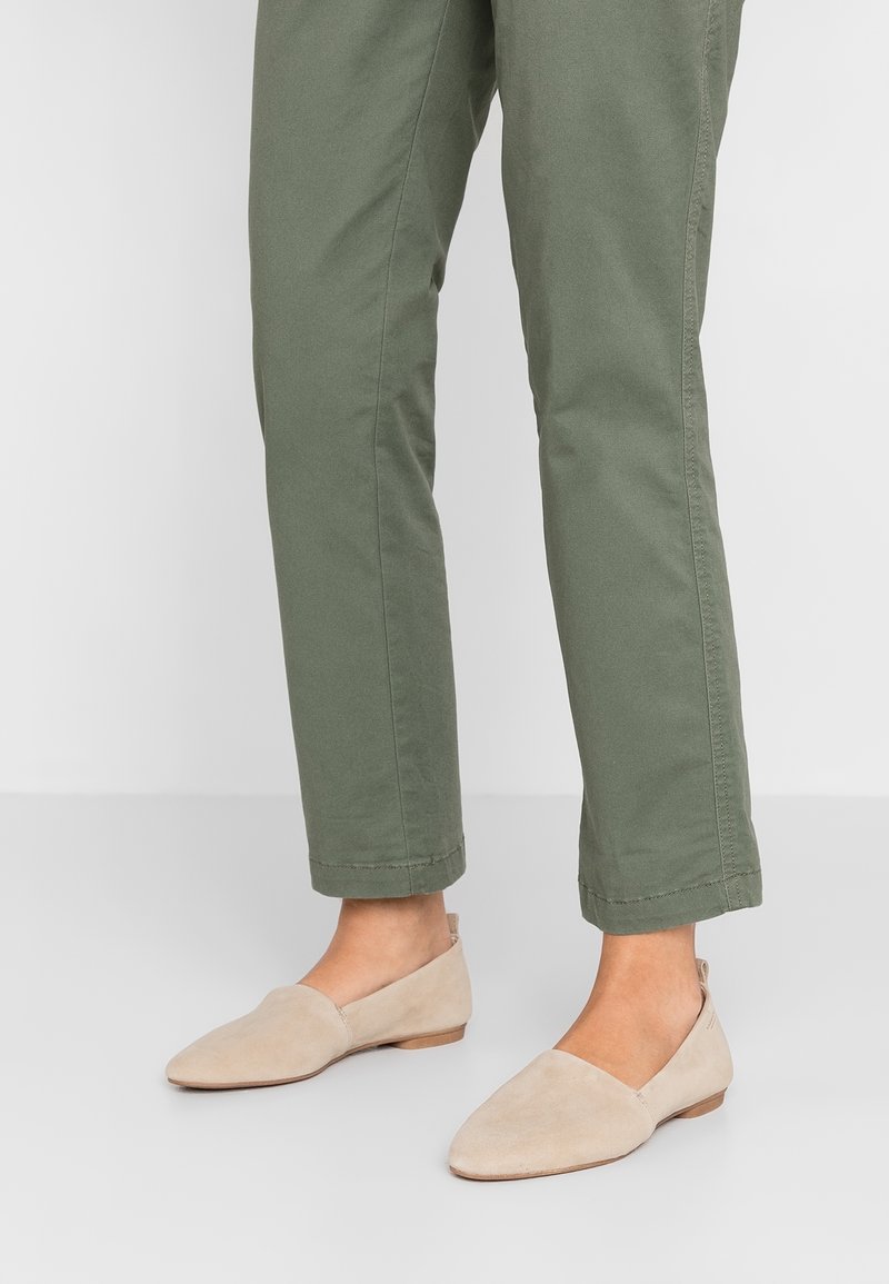 Personne portant un pantalon court vert olive et des chaussures plates beige à enfiler, debout sur un sol gris clair.