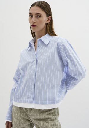 Chemise boutonnée bleu clair et blanche à rayures, avec un col ouvert et des épaules tombantes, associée à un pantalon taille haute texturé de couleur vert clair.