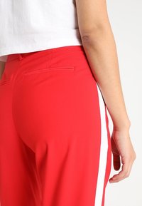 Rote Hosen mit weißen Seitenstreifen, die eine glatte Textur und einen schmalen Schnitt aufweisen. Auf der Rückseite befinden sich zwei eingesetzte Taschen. Modell von der Seite gezeigt.
