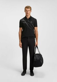 Zwarte polo shirt met een witte lijnontwerp, gecombineerd met zwarte broek en een zwarte sporttas. Vloeiende stof, korte mouwen en kraag.