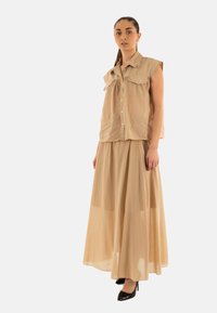 Blusa sin mangas beige con bolsillos frontales y cierre de botones, combinada con una falda maxi fluida a juego, textura suave y silueta ajustada.