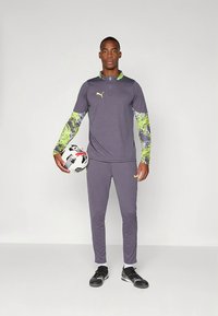 Puma sportinė apranga pilka spalva su ryškiomis žaliomis ir violetinėmis raštuotomis rankovėmis; modelis laiko futbolo kamuolį, dėvi juodus batus.