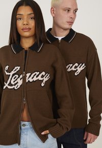 Pull en maille marron zippé avec col noir, arborant un texte "Legacy" en blanc et rose dans un style cursif. Poignets et ourlet côtelés.