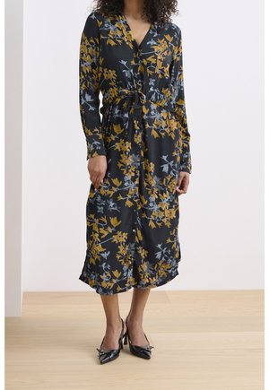 Robe noire à motifs floraux jaunes et bleus, avec un front à boutons, des manches longues, une taille cintrée et des poches latérales. Associée à des talons noirs.