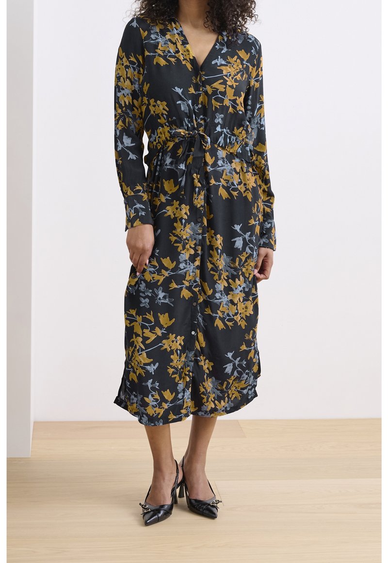 Robe noire à motifs floraux jaunes et bleus, avec un front à boutons, des manches longues, une taille cintrée et des poches latérales. Associée à des talons noirs.