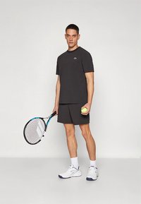 Svart trenings-T-skjorte og shorts, hvite joggesko, holder en tennisracket og ball. Enkelt design med en liten logo på skjorten.
