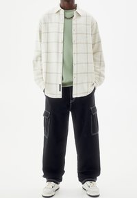 Camisa de cuadros blanca con botones sobre un suéter verde; combinada con pantalones cargo negros con bolsillos y zapatillas blancas con detalles en negro.