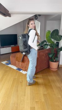 Musta, kestävä reppu, jossa on säädettävät hihnat, sivutaskut ja suuri logo. Kannan henkilö, jolla on pitkät hiukset, modernin asuintilan taustalla.