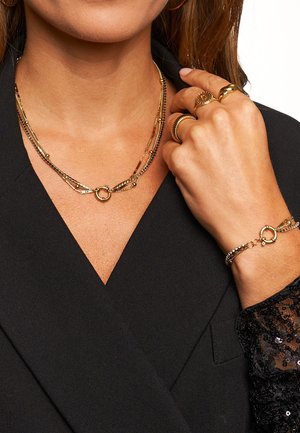 Gouden gelaagde ketting met een ronde sluiting, gecombineerd met een dunne gouden armband met decoratieve accenten. Getextureerde zwarte jas op de achtergrond.