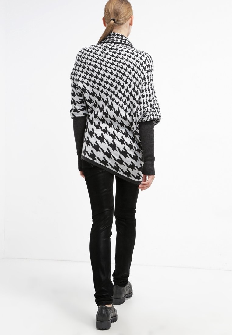 Femme vue de dos portant un châle pied-de-poule noir et blanc, un haut à manches longues noir, un pantalon noir et des chaussures grises sur fond blanc.