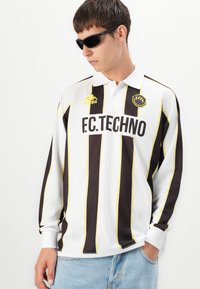 Hvit langermet polo med svarte og gule vertikale striper. Inkluderer "F.C.TECHNO"-logo og "Kappa"-merke. Brukt med mørke solbriller.