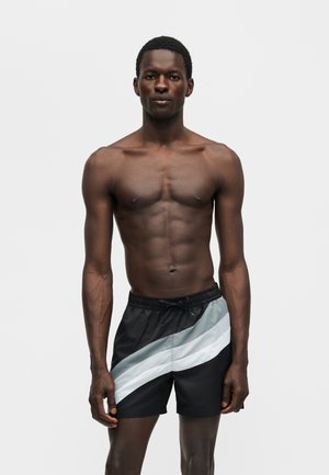 Αθλητικός μαύρος άνδρας στέκεται ακίνητος, φορώντας μαύρο μαγιό με δύο διαγώνιες γκρι και λευκές ρίγες σε μονόχρωμο ανοιχτό φόντο.