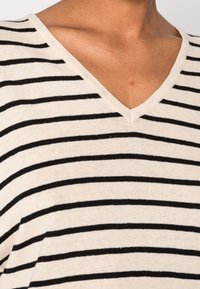 V-Ausschnitt Pullover in Creme und schwarzen Streifen, mit weicher Textur und lockerem Schnitt. Der Stoff wirkt leicht und atmungsaktiv.