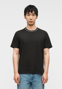 Μαύρο t-shirt με διακριτικό μοτίβο λογότυπου, κοντά μανίκια και στρογγυλή λαιμόκοψη με λευκή επένδυση, συνδυασμένο με μπλε τζιν παντελόνι.