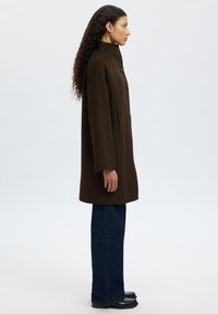 Manteau en mélange de laine marron avec un col haut, des boutons sur le devant et des poches latérales, associé à un jean ample foncé et des chaussures noires.