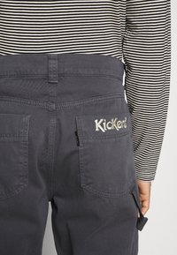 Personne portant un pantalon gris foncé avec « Kickers » brodé sur la poche arrière et un t-shirt à manches longues rayé noir et blanc.