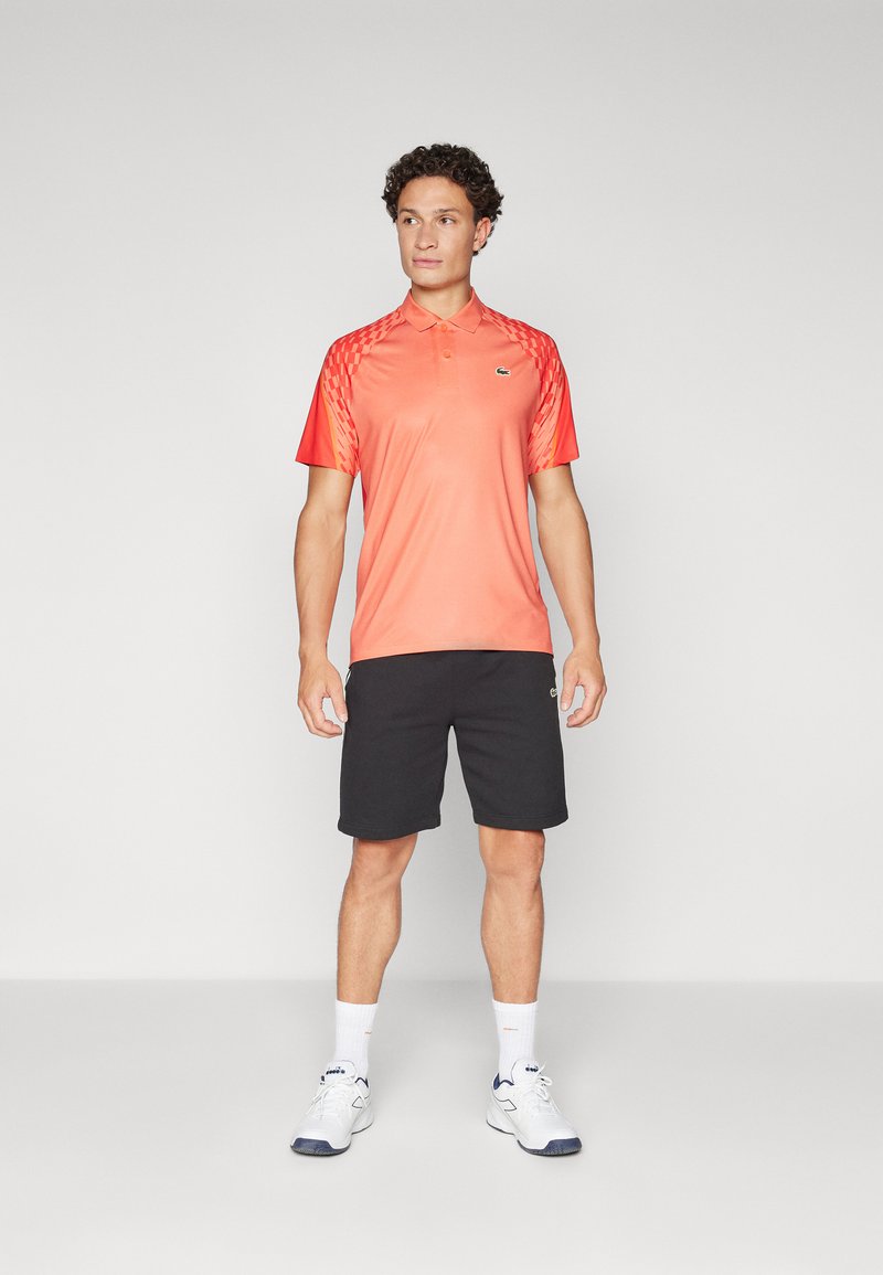 Herren Poloshirt in leuchtendem Koralle mit kurzen Ärmeln, das ein strukturiertes Verlaufsdesign zeigt, kombiniert mit schwarzen Shorts und weißen Sneakers.